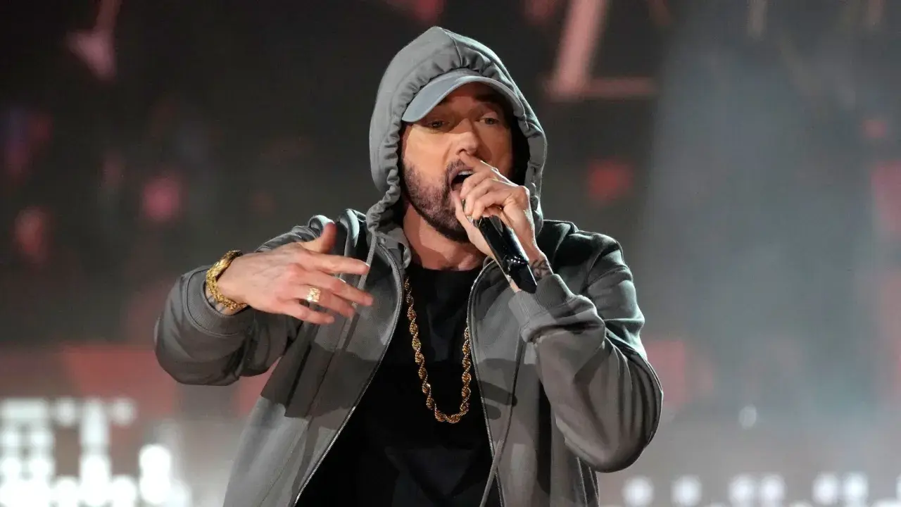 Eminem w szarej bluzie z kapturem i złotym łańcuchem śpiewa do mikrofonu.