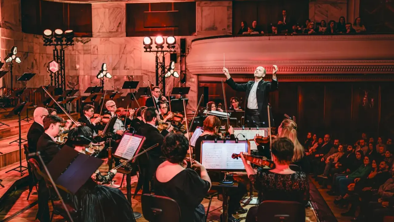 Dyrygent prowadzi orkiestrę filharmoniczną. Koncert trwa, a widzowie na balkonie wsłuchują się w muzykę.