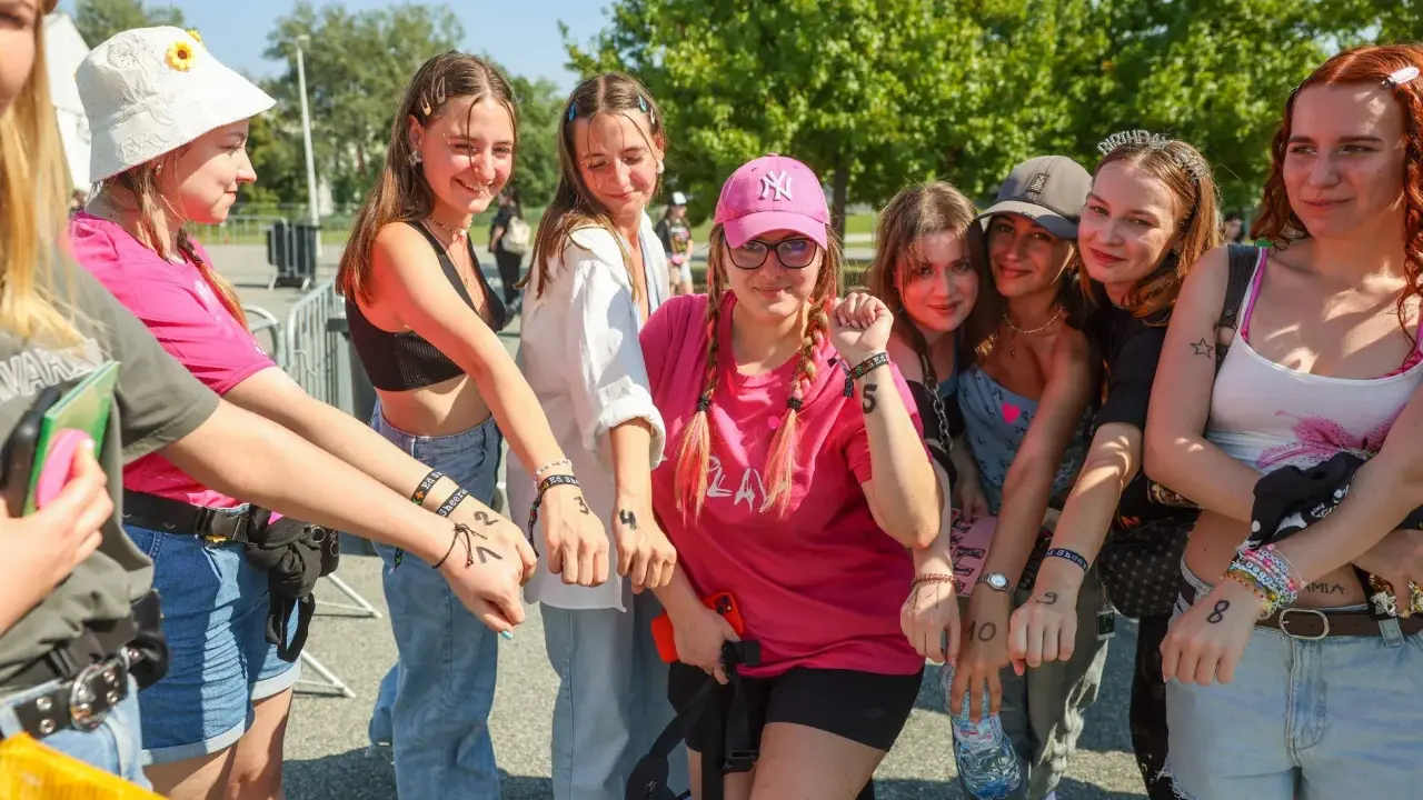 Grupa młodych kobiet z numerami na rękach, podekscytowana przed koncertem. Czekają, by dowiedzieć się, ile trwa koncert Eda Sheerana.