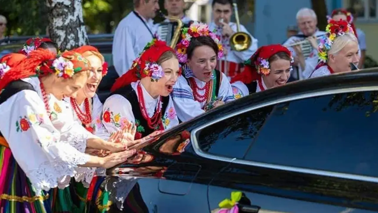 Kobiety w strojach ludowych śpiewają, jak w tekście piosenki "Gdzie ci mężczyźni", przy czarnym samochodzie ozdobionym wstążkami.