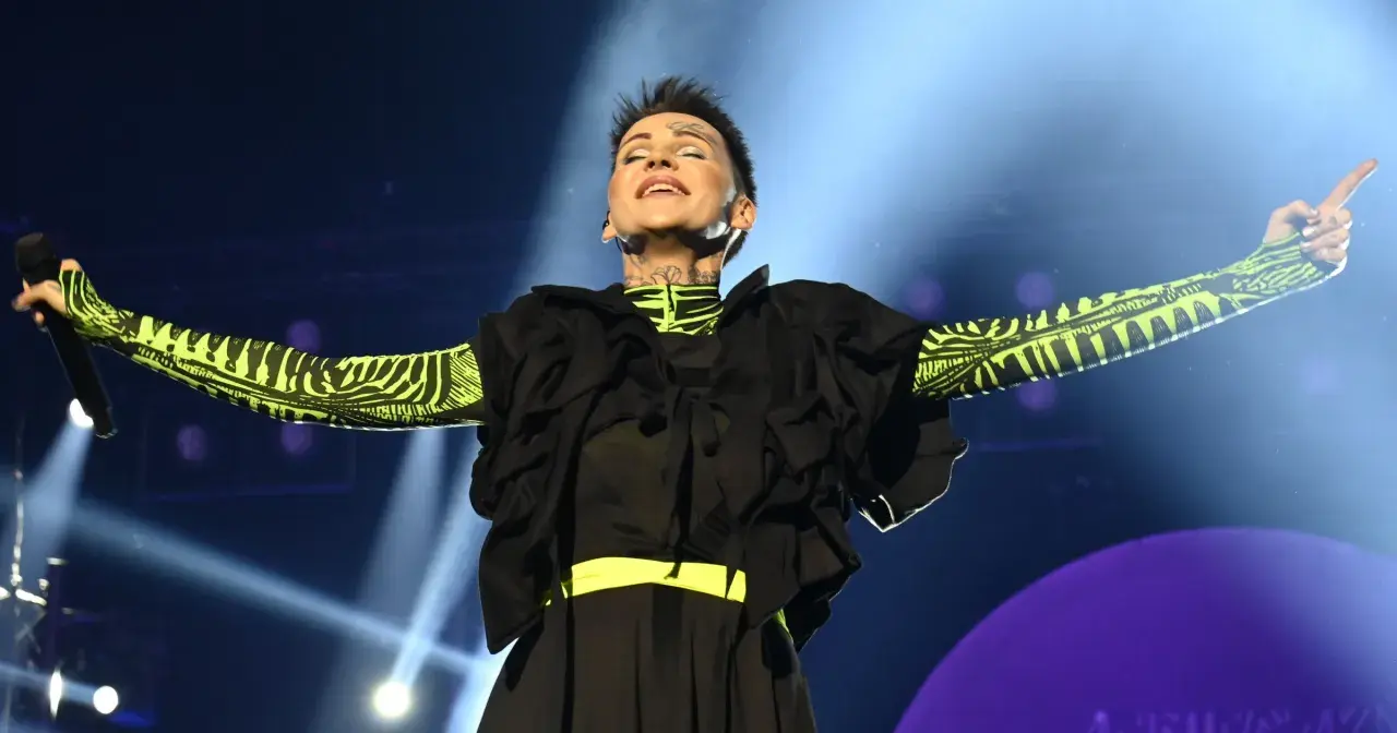 Chylińska na scenie, z mikrofonem w dłoni, w czarnej sukni i bluzie z neonowym wzorem. Fani zastanawiają się, ile trwa koncert Chylińskiej.