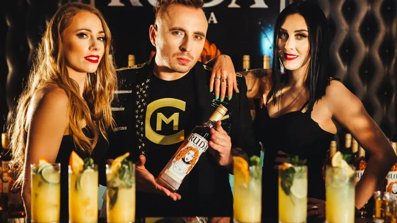 Trzy osoby pozują z butelką wódki Ruda. W tle widać drinki i logo.