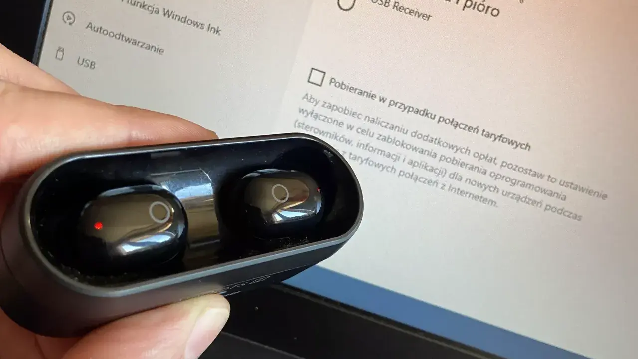 Dłoń trzyma czarne słuchawki w etui, obok ekranu laptopa z ustawieniami, pokazując jak sparować słuchawki z laptopem.