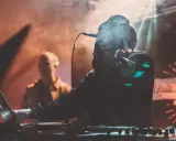 DJ w dymie, z mikrofonem, tworzy muzykę. W tle widać drugą osobę. To jak tekst piosenki "Dni których nie znamy".