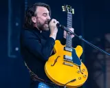 Mężczyzna z brodą i kręconymi włosami śpiewa do mikrofonu, trzymając żółtą gitarę. Czuje, że "kto śpiewa mój jest ten kawałek podłogi".