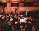 Dyrygent prowadzi orkiestrę filharmoniczną. Koncert trwa, a widzowie na balkonie wsłuchują się w muzykę.