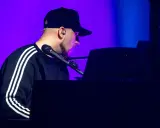Artysta w czapce z daszkiem gra na pianinie. Koncert Korteza trwa, a fioletowe światło podkreśla atmosferę.