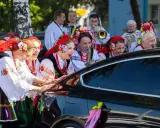 Kobiety w strojach ludowych śpiewają, jak w tekście piosenki "Gdzie ci mężczyźni", przy czarnym samochodzie ozdobionym wstążkami.