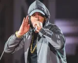 Eminem w szarej bluzie z kapturem i czapce, z mikrofonem w dłoni. Choć nie widać tego na zdjęciu, eminem ile ma wzrostu to 178 cm.