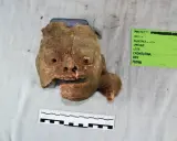 Fragment glinianej maski, jak z tekstu piosenki Kaen "Kim jestem?", z widocznymi otworami na oczy i usta, obok zielonej etykiety z datą 23/08/2018.