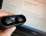 Dłoń trzyma czarne słuchawki w etui, obok ekranu laptopa z ustawieniami, pokazując jak sparować słuchawki z laptopem.