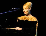 Kobieta w czarnej sukni z ozdobnym kołnierzem gra na instrumencie, tworząc muzykę. Kto śpiewa?