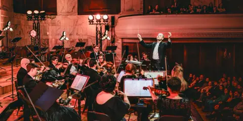 Dyrygent prowadzi orkiestrę filharmoniczną. Koncert trwa, a widzowie na balkonie wsłuchują się w muzykę.