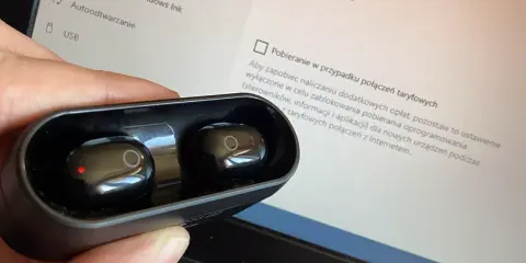 Dłoń trzyma czarne słuchawki w etui, obok ekranu laptopa z ustawieniami, pokazując jak sparować słuchawki z laptopem.