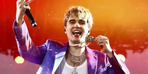 Raper Mata w błyszczącej marynarce, z mikrofonem i naszyjnikiem z napisem "Mata". Zastanawiasz się, ile kosztuje bilet na koncert Maty?