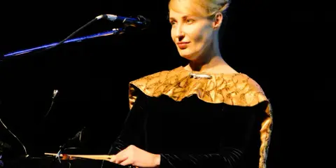 Kobieta w czarnej sukni z ozdobnym kołnierzem gra na instrumencie, tworząc muzykę. Kto śpiewa?