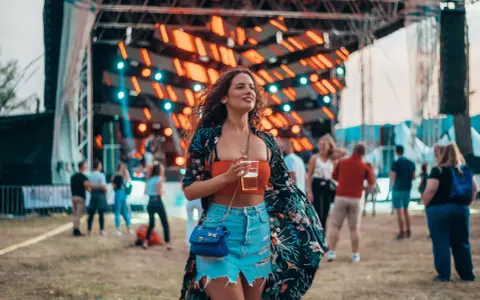 Dziewczyna w kwiecistej narzutce i jeansowej spódniczce, z drinkiem w ręku, pokazuje, jak ubrać się na koncert.