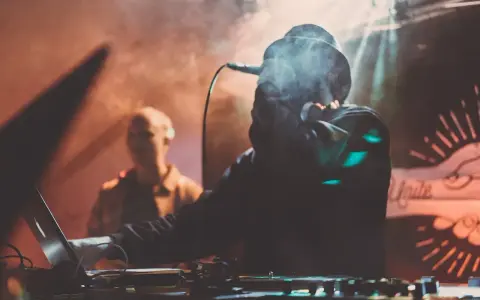 DJ w dymie, z mikrofonem, tworzy muzykę. W tle widać drugą osobę. To jak tekst piosenki "Dni których nie znamy".