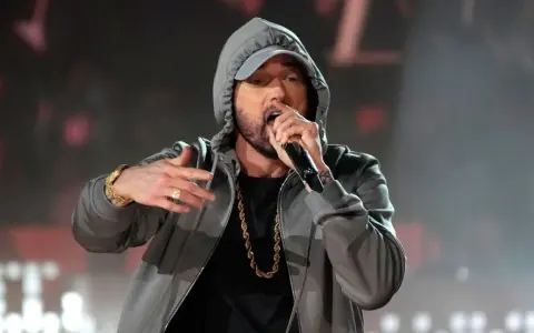 Eminem w szarej bluzie z kapturem i złotym łańcuchem śpiewa do mikrofonu.