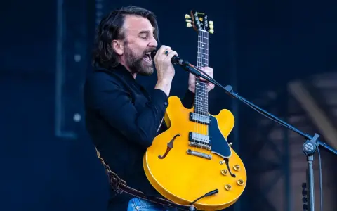 Mężczyzna z brodą i kręconymi włosami śpiewa do mikrofonu, trzymając żółtą gitarę. Czuje, że "kto śpiewa mój jest ten kawałek podłogi".