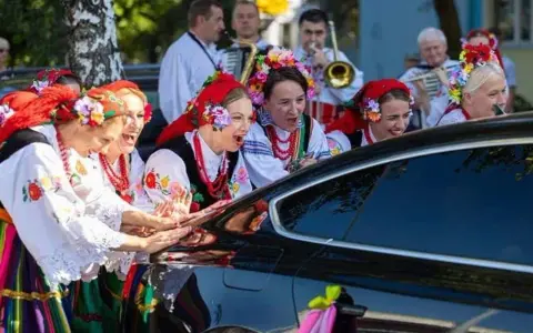 Kobiety w strojach ludowych śpiewają, jak w tekście piosenki "Gdzie ci mężczyźni", przy czarnym samochodzie ozdobionym wstążkami.