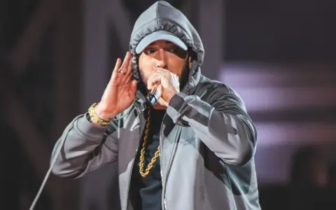 Eminem w szarej bluzie z kapturem i czapce, z mikrofonem w dłoni. Choć nie widać tego na zdjęciu, eminem ile ma wzrostu to 178 cm.