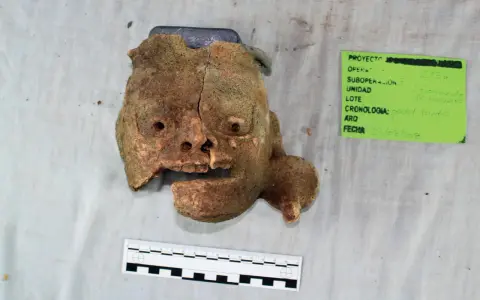 Fragment glinianej maski, jak z tekstu piosenki Kaen "Kim jestem?", z widocznymi otworami na oczy i usta, obok zielonej etykiety z datą 23/08/2018.