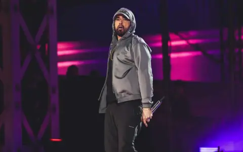 Eminem na scenie w szarej bluzie z kapturem i czarnych spodniach, trzymający mikrofon.