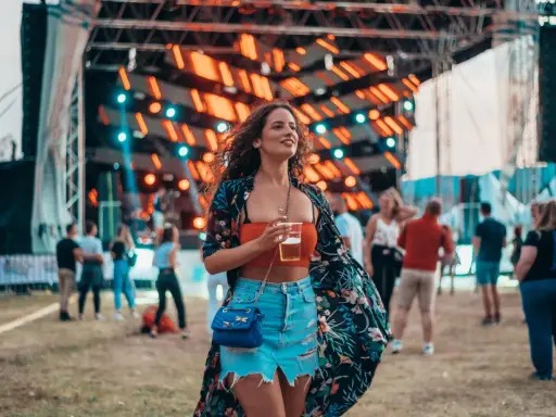 Dziewczyna w kwiecistej narzutce i jeansowej spódniczce, z drinkiem w ręku, pokazuje, jak ubrać się na koncert.