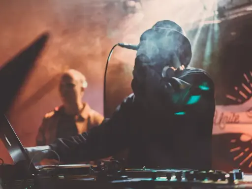 DJ w dymie, z mikrofonem, tworzy muzykę. W tle widać drugą osobę. To jak tekst piosenki "Dni których nie znamy".