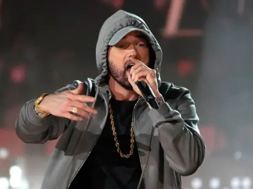 Eminem w szarej bluzie z kapturem i złotym łańcuchem śpiewa do mikrofonu.
