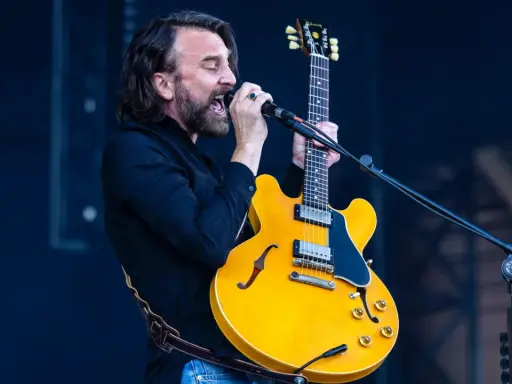 Mężczyzna z brodą i kręconymi włosami śpiewa do mikrofonu, trzymając żółtą gitarę. Czuje, że "kto śpiewa mój jest ten kawałek podłogi".