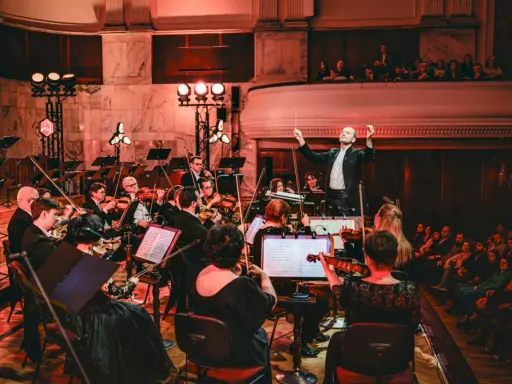Dyrygent prowadzi orkiestrę filharmoniczną. Koncert trwa, a widzowie na balkonie wsłuchują się w muzykę.