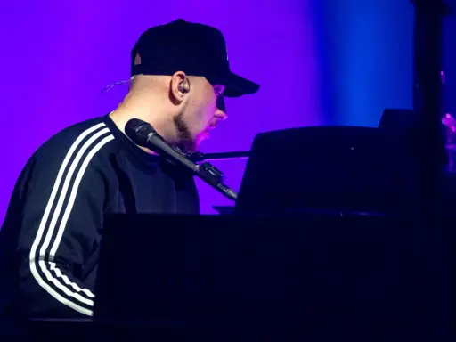Artysta w czapce z daszkiem gra na pianinie. Koncert Korteza trwa, a fioletowe światło podkreśla atmosferę.