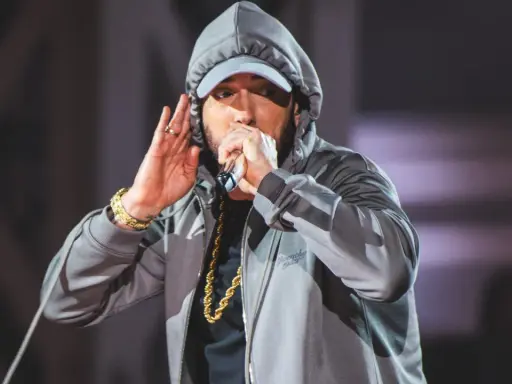 Eminem w szarej bluzie z kapturem i czapce, z mikrofonem w dłoni. Choć nie widać tego na zdjęciu, eminem ile ma wzrostu to 178 cm.