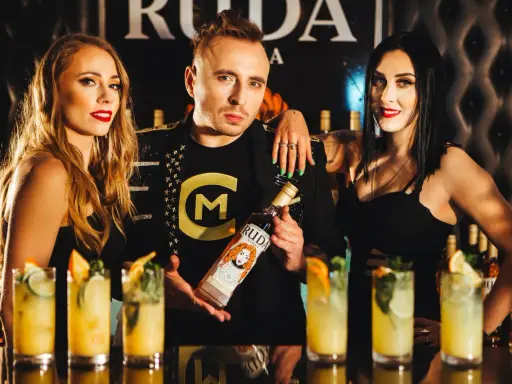 Trzy osoby pozują z butelką wódki Ruda. W tle widać drinki i logo.
