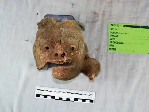 Fragment glinianej maski, jak z tekstu piosenki Kaen "Kim jestem?", z widocznymi otworami na oczy i usta, obok zielonej etykiety z datą 23/08/2018.