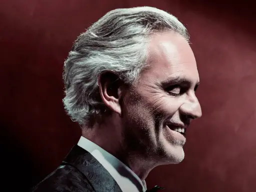 Uśmiechnięty Andrea Bocelli w garniturze, z siwymi włosami, w profilu. To zapowiedź, gdzie koncert Bocellego.