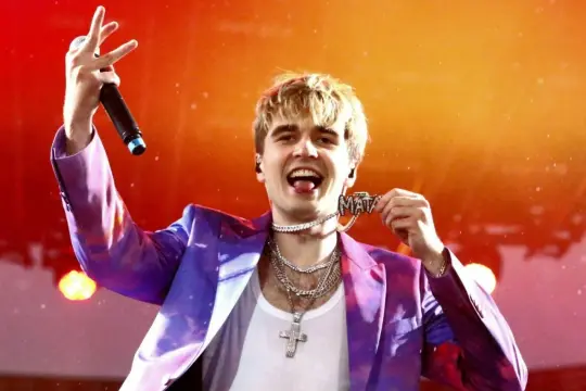 Raper Mata w błyszczącej marynarce, z mikrofonem i naszyjnikiem z napisem "Mata". Zastanawiasz się, ile kosztuje bilet na koncert Maty?