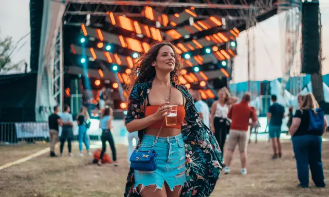 Dziewczyna w kwiecistej narzutce i jeansowej spódniczce, z drinkiem w ręku, pokazuje, jak ubrać się na koncert.