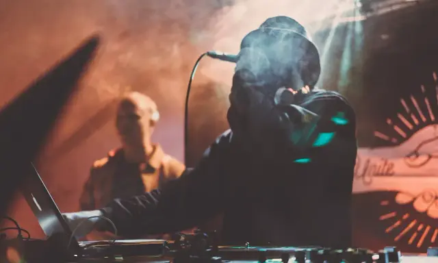 DJ w dymie, z mikrofonem, tworzy muzykę. W tle widać drugą osobę. To jak tekst piosenki "Dni których nie znamy".
