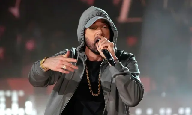 Eminem w szarej bluzie z kapturem i złotym łańcuchem śpiewa do mikrofonu.