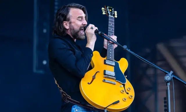 Mężczyzna z brodą i kręconymi włosami śpiewa do mikrofonu, trzymając żółtą gitarę. Czuje, że "kto śpiewa mój jest ten kawałek podłogi".