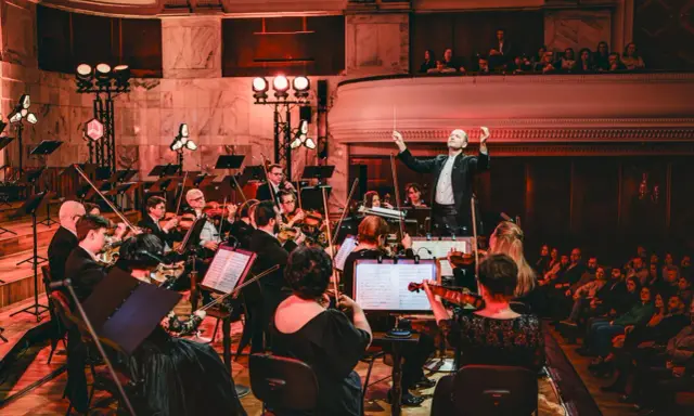 Dyrygent prowadzi orkiestrę filharmoniczną. Koncert trwa, a widzowie na balkonie wsłuchują się w muzykę.