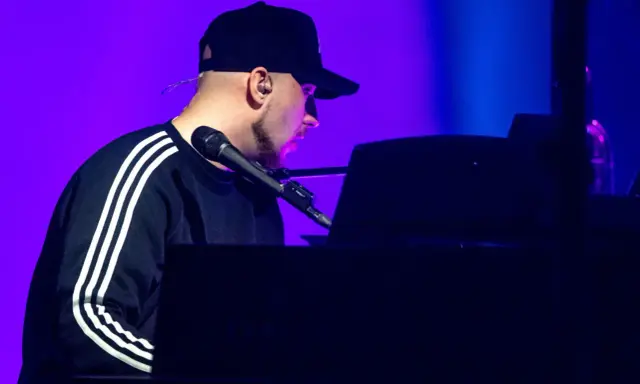 Artysta w czapce z daszkiem gra na pianinie. Koncert Korteza trwa, a fioletowe światło podkreśla atmosferę.
