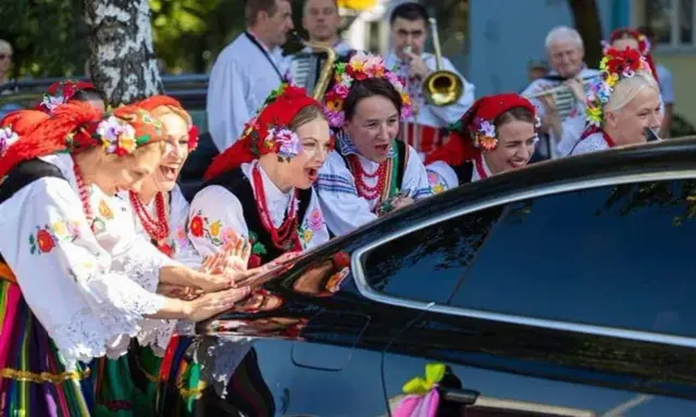 Kobiety w strojach ludowych śpiewają, jak w tekście piosenki "Gdzie ci mężczyźni", przy czarnym samochodzie ozdobionym wstążkami.