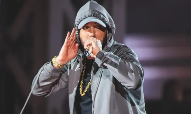 Eminem w szarej bluzie z kapturem i czapce, z mikrofonem w dłoni. Choć nie widać tego na zdjęciu, eminem ile ma wzrostu to 178 cm.