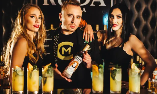 Trzy osoby pozują z butelką wódki Ruda. W tle widać drinki i logo.