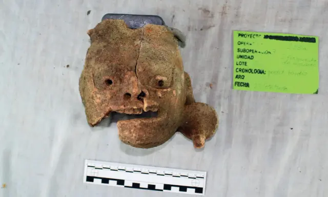 Fragment glinianej maski, jak z tekstu piosenki Kaen "Kim jestem?", z widocznymi otworami na oczy i usta, obok zielonej etykiety z datą 23/08/2018.