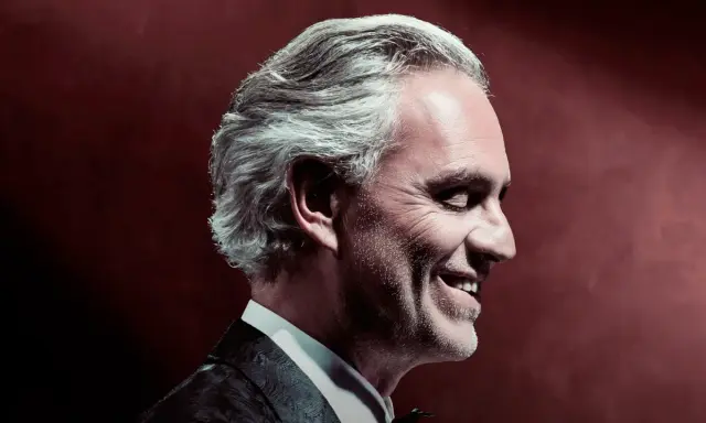 Uśmiechnięty Andrea Bocelli w garniturze, z siwymi włosami, w profilu. To zapowiedź, gdzie koncert Bocellego.